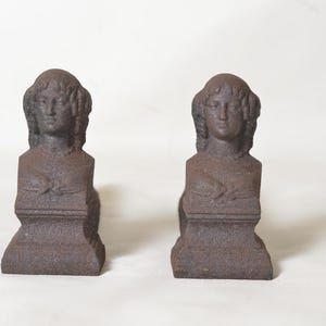 Pode incluir: Um par de suportes para livros de ferro fundido texturizado em castanho escuro, cada um apresentando o busto esculpido de uma pessoa com cabelo ondulado, um colar e uma peça de vestuário drapeada, sobre uma base retangular escalonada.