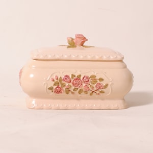 Peut inclure: Une boîte à bijoux en céramique blanche avec un motif floral composé de roses roses et de feuilles vertes. Le couvercle est surmonté d'une rose rose en relief.