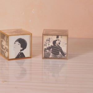 Op de afbeelding: Twee transparante glazen fotolijsten met zwart-witfoto's van kinderen. De lijsten zijn kubusvormig en hebben een lichtbruine tint.