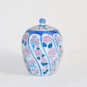 Könnte beinhalten: Eine weiße Keramikdose mit Deckel, verziert mit rosa und blauen Blumendesigns. Die Dose hat eine abgerundete Form und eine leicht ausgestellte Öffnung.