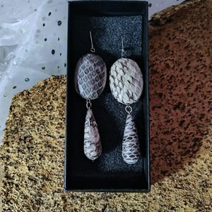 Könnte beinhalten: Ein Paar Hängeohrringe mit einem tropfenförmigen Anhänger. Die Ohrringe sind aus Silber und haben ein graues und weißes Schlangenhautmuster.