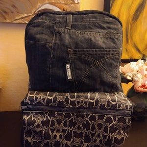 Könnte beinhalten: Eine schwarze und graue Clutch-Tasche mit Schlangenhautmuster und Reißverschluss, gestapelt auf einem dunkelblauen Jeans-Rucksack mit einem Etikett mit der Aufschrift "C. Grande".