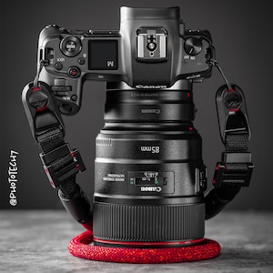 May include: A black Canon camera with an 85mm lens attached. The camera has a black strap with red accents. The lens has a red ring around the front.