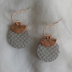 May include: A pair of dangle earrings with a gold-colored hook and a half-circle accent. The earrings feature a gray, textured, circular pendant with a scale-like pattern. The earrings are displayed on a white, textured surface.