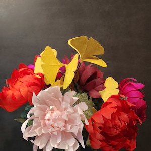 May include: A bouquet of paper flowers in a glass vase. The flowers are red, pink, and burgundy, with yellow ginkgo leaves.