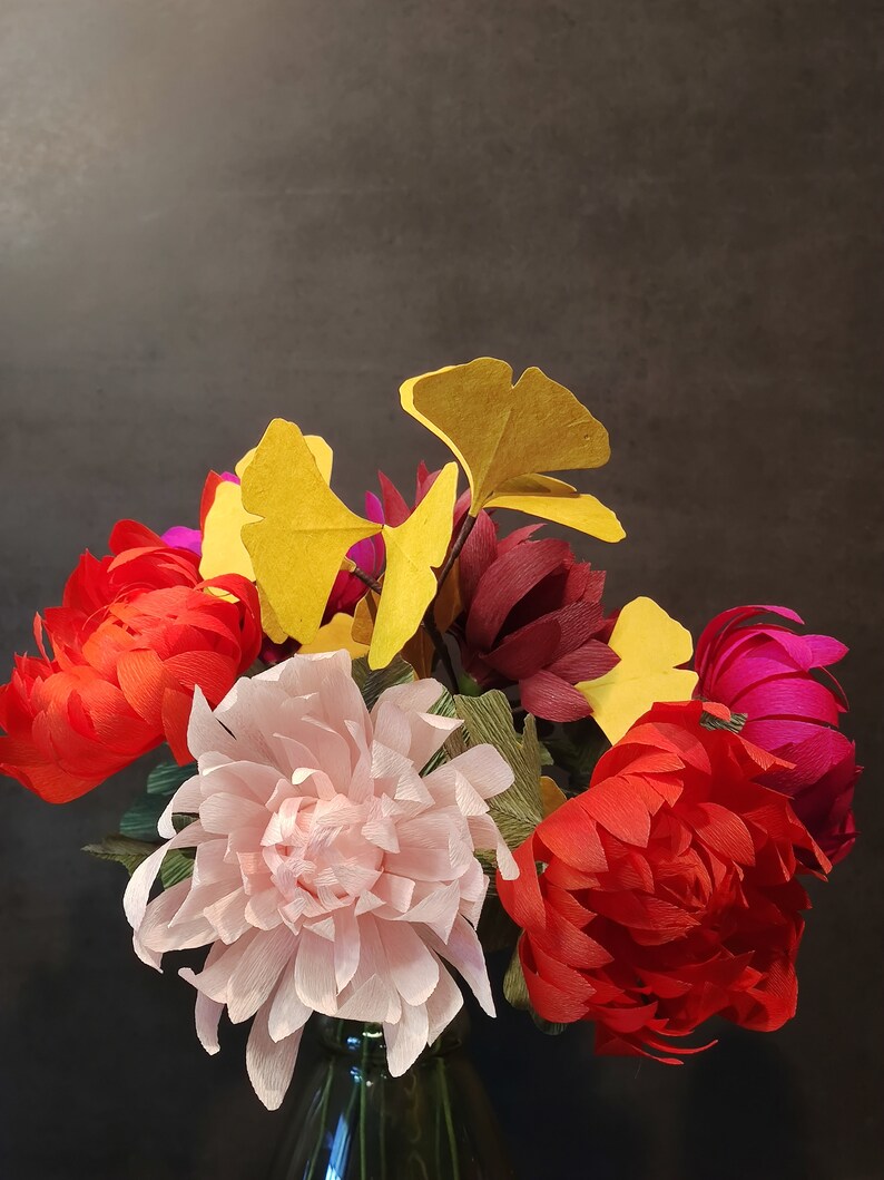 May include: A bouquet of paper flowers in a glass vase. The flowers are red, pink, and burgundy, with yellow ginkgo leaves.