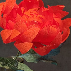 May include: A vibrant red paper flower with crinkled petals, arranged in a vase with green leaves.