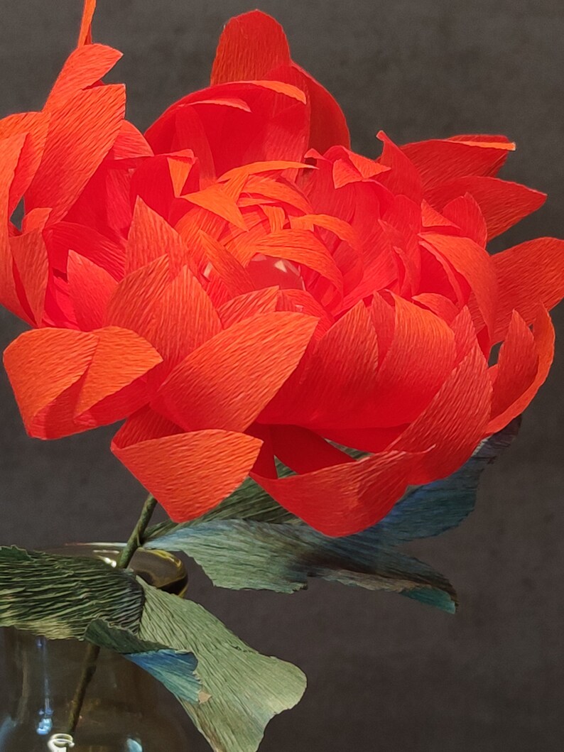 May include: A vibrant red paper flower with crinkled petals, arranged in a vase with green leaves.