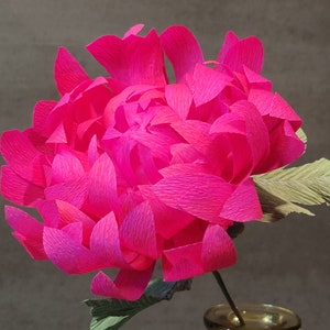 May include: A vibrant pink paper flower with green leaves in a clear glass vase. The flower is made of multiple layers of crinkled paper, creating a full and textured appearance.