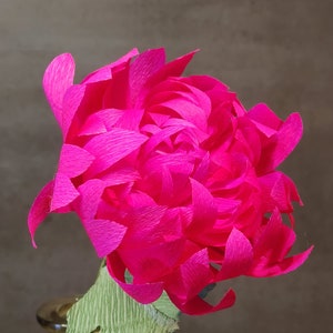 May include: A vibrant pink paper flower with a green paper stem. The flower is made of many layers of paper, giving it a full and fluffy appearance.