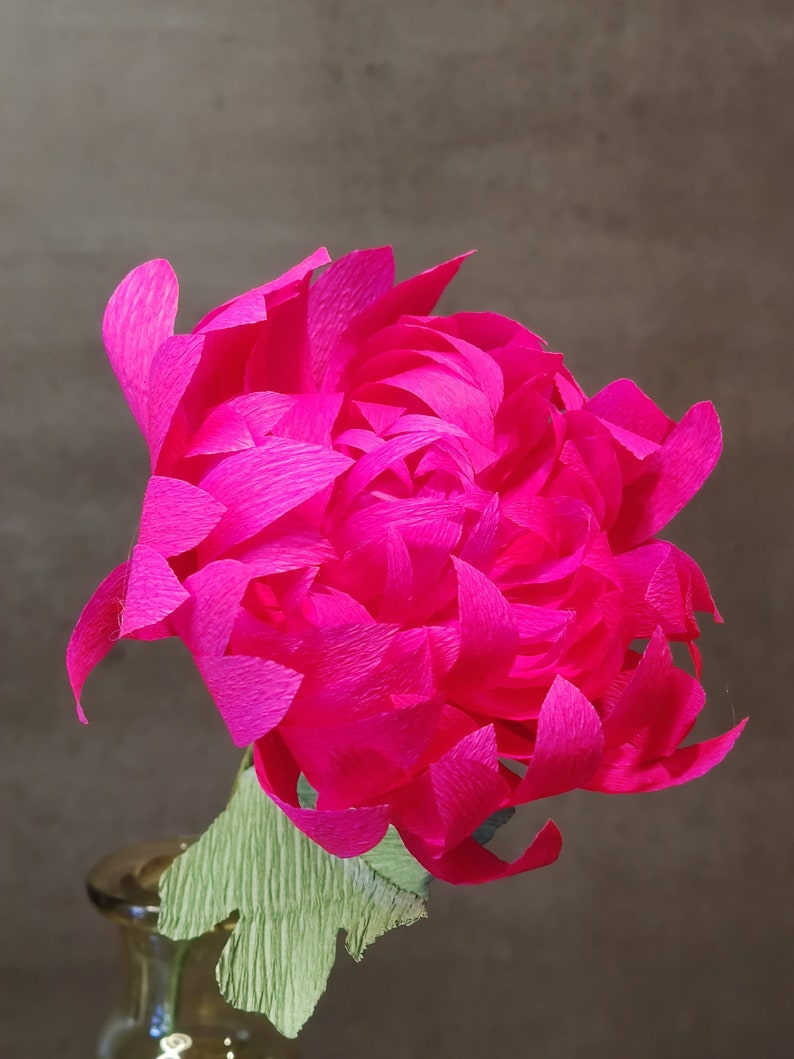 May include: A vibrant pink paper flower with a green paper stem. The flower is made of many layers of paper, giving it a full and fluffy appearance.