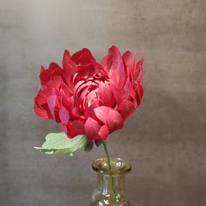 May include: A single red paper flower with green leaves in a clear glass vase. The vase has a rounded bottom and a wide mouth.