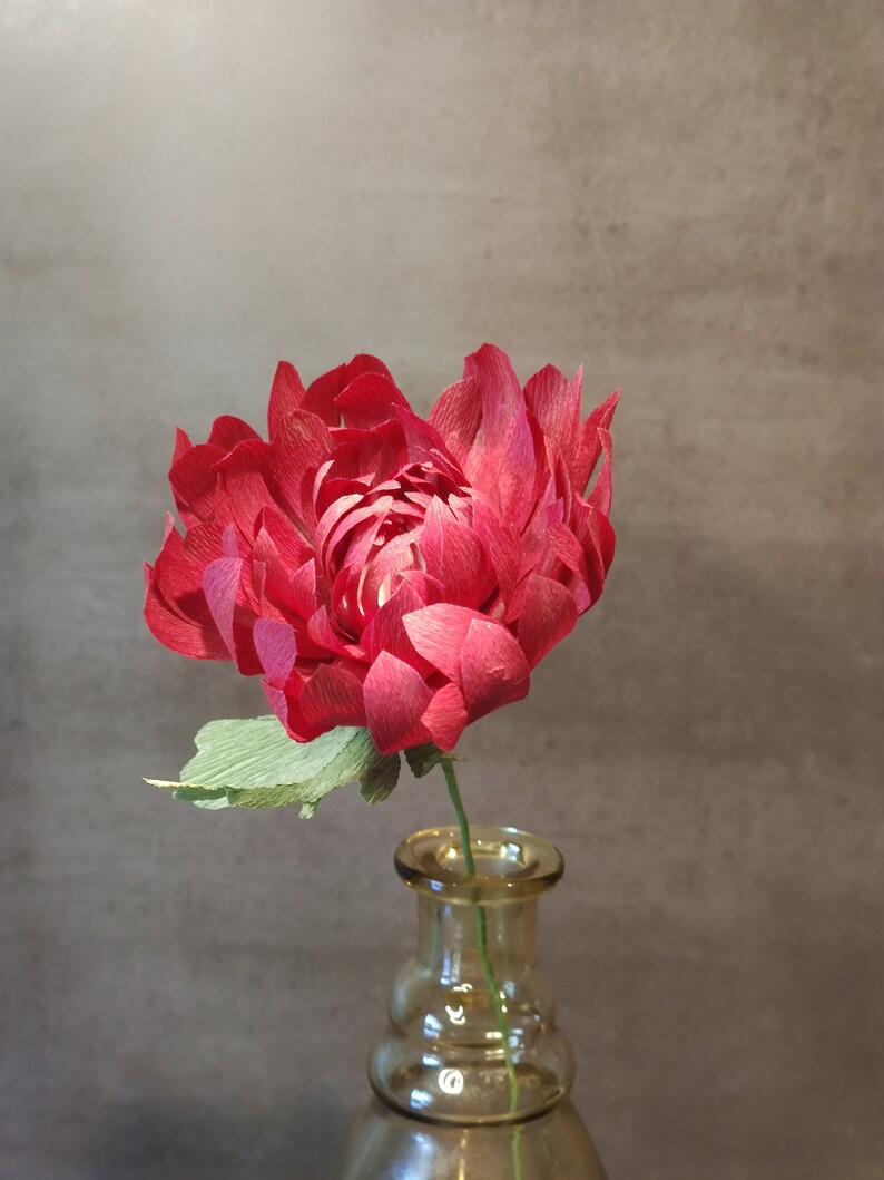 May include: A single red paper flower with green leaves in a clear glass vase. The vase has a rounded bottom and a wide mouth.