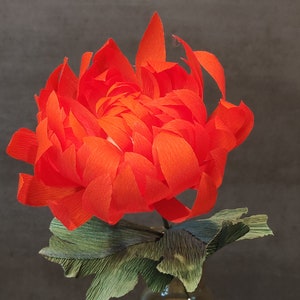 May include: A vibrant orange paper flower with green paper leaves in a clear glass vase. The flower is made of crinkled paper and has a delicate, layered appearance.