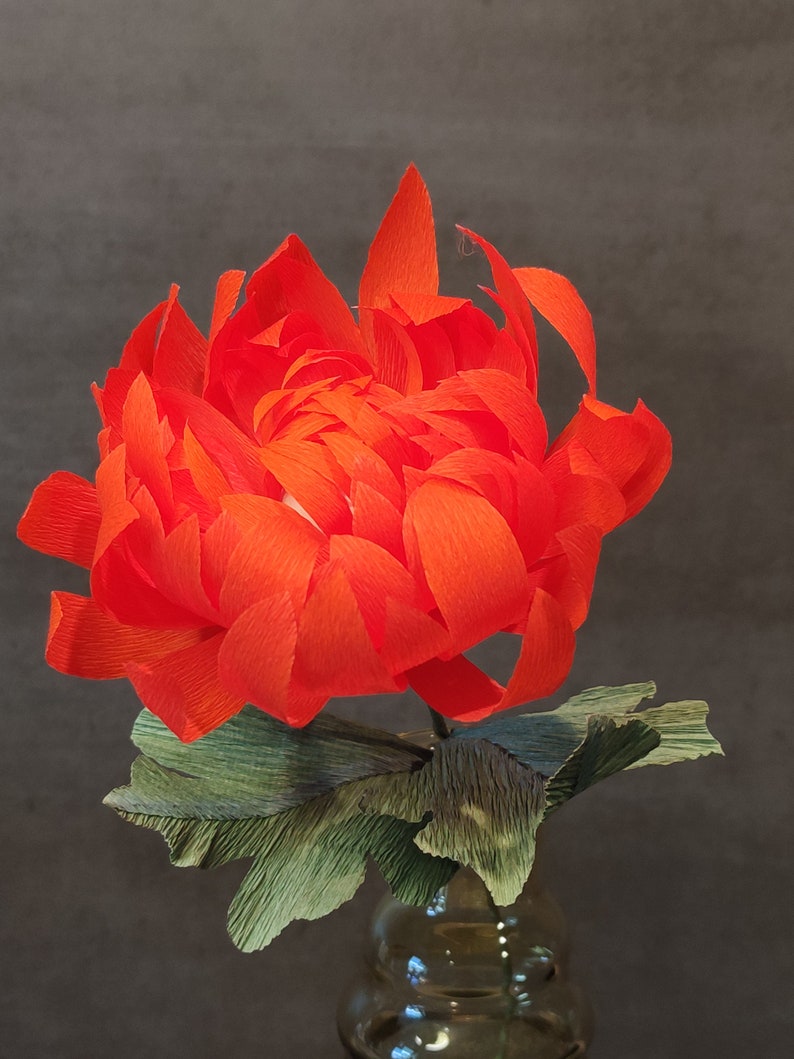 May include: A vibrant orange paper flower with green paper leaves in a clear glass vase. The flower is made of crinkled paper and has a delicate, layered appearance.