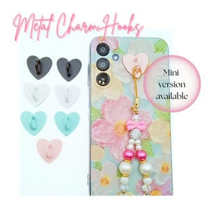 May include: A collection of heart-shaped metal charm hooks in black, white, teal, and pink. A floral phone case with a pink heart charm and beaded lanyard is also shown. Text reads "Metal Charm Hooks" and "Mini version available."