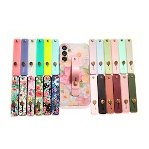 May include: A collection of rectangular phone finger straps in various solid colors and floral patterns, arranged around a central smartphone case featuring a matching floral design and a pink finger strap attached to the back of the device.