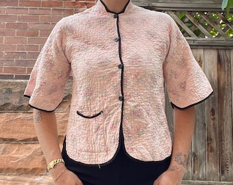 May include: A pink floral quilted jacket with black trim and a mandarin collar. The jacket has a button closure and short sleeves.