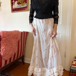 May include: A black lace long-sleeve top paired with a white floral print skirt. The skirt has a ruffled hem. The outfit is vintage-inspired and suitable for a special occasion or everyday wear.