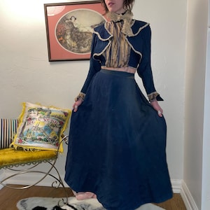 May include: A woman wearing a dark blue long skirt with a matching cropped jacket featuring lace trim and a striped blouse underneath. The jacket has a large, ruffled collar.