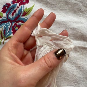 May include: Close-up of a section of white fabric with a ruffled edge, held by a hand. The fabric has a delicate, sheer appearance. The background is a floral patterned cloth with red, blue, and green elements.