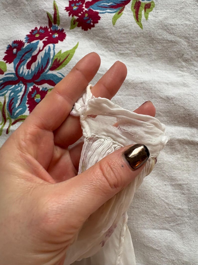 May include: Close-up of a section of white fabric with a ruffled edge, held by a hand. The fabric has a delicate, sheer appearance. The background is a floral patterned cloth with red, blue, and green elements.
