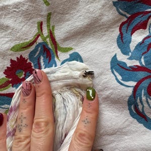 May include: Close-up of a hand with painted nails holding a white fabric with a metal clip. The background is a white floral patterned fabric with blue, red, and green accents.
