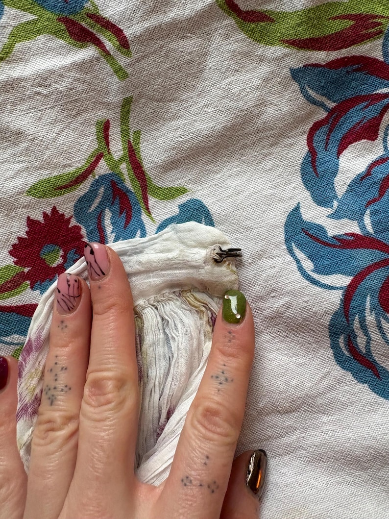 May include: Close-up of a hand with painted nails holding a white fabric with a metal clip. The background is a white floral patterned fabric with blue, red, and green accents.