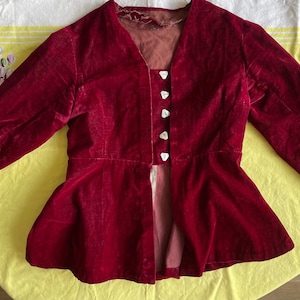 May include: A deep red velvet jacket with a V-neck and long sleeves. The jacket features a peplum waist and is fastened with four white buttons. The garment is displayed on a yellow floral tablecloth.