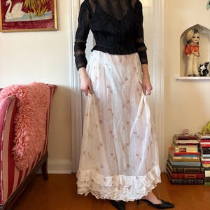 May include: A black long-sleeved top with a sheer, textured design is paired with a long, white skirt adorned with small pink floral patterns. The skirt features a ruffled hemline. The outfit is worn with black shoes.