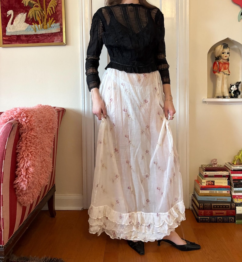 May include: A black long-sleeved top with a sheer, textured design is paired with a long, white skirt adorned with small pink floral patterns. The skirt features a ruffled hemline. The outfit is worn with black shoes.