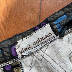 Op de afbeelding: Close-up van een zwarte en gouden stof met een wit label waarop "alex colman california" staat.