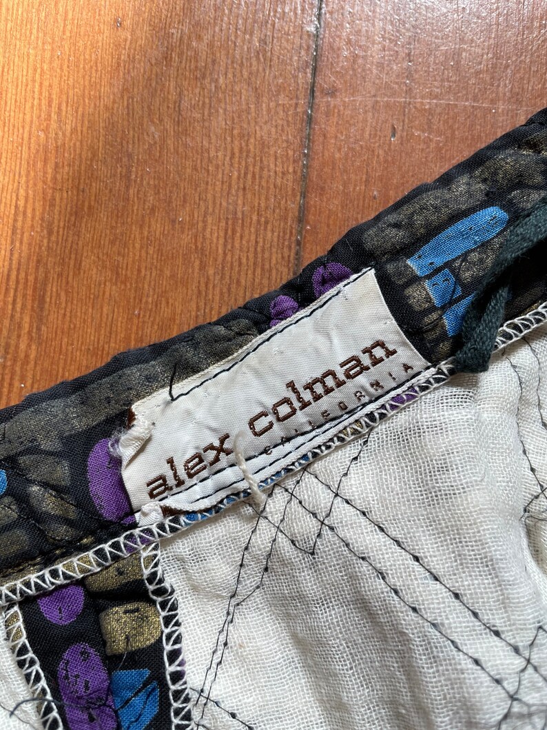 Op de afbeelding: Close-up van een zwarte en gouden stof met een wit label waarop "alex colman california" staat.