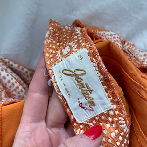 Pu&ograve; includere: Tessuto a fantasia arancione e bianca con un'etichetta dorata "Jantzen" che recita "Made in USA" e un'etichetta di taglia "12".