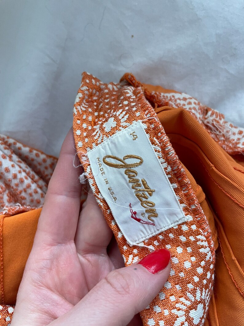 Pu&ograve; includere: Tessuto a fantasia arancione e bianca con un'etichetta dorata "Jantzen" che recita "Made in USA" e un'etichetta di taglia "12".