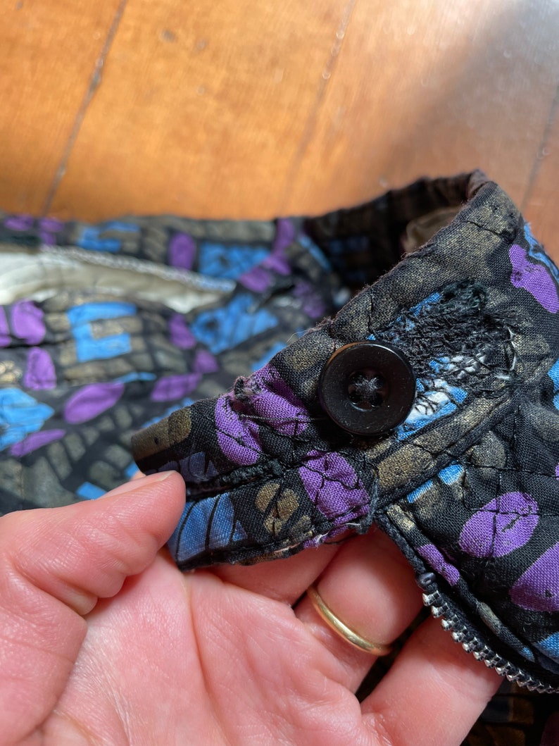 Op de afbeelding: Close-up van een zwart gewatteerd jack met een paarse en blauwe bloemenprint. Het jack heeft een ritssluiting en een enkele zwarte knoop.