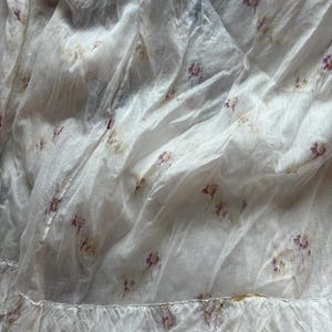 May include: White fabric with a delicate floral pattern in shades of purple and yellow. The fabric has a ruffled edge, adding a touch of elegance. The material appears lightweight and slightly translucent, suggesting a soft texture.