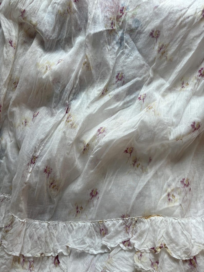May include: White fabric with a delicate floral pattern in shades of purple and yellow. The fabric has a ruffled edge, adding a touch of elegance. The material appears lightweight and slightly translucent, suggesting a soft texture.
