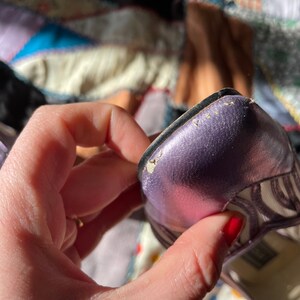 Pu&ograve; includere: Primo piano di una scarpa con tacco aperto a punta aperta di colore viola metallizzato con un motivo a righe bianche e nere. La scarpa presenta un leggero graffio sulla punta.