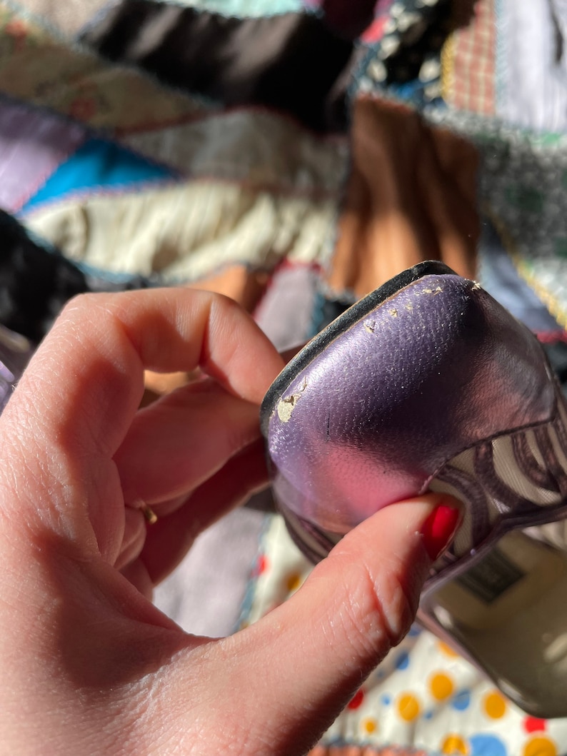 Pu&ograve; includere: Primo piano di una scarpa con tacco aperto a punta aperta di colore viola metallizzato con un motivo a righe bianche e nere. La scarpa presenta un leggero graffio sulla punta.