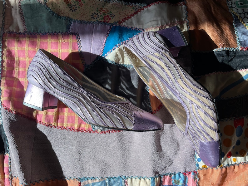 Pu&ograve; includere: Un paio di scarpe con tacco in rete viola con un motivo ondulato. Le scarpe hanno un tacco a blocco basso e una punta a punta.