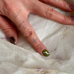 May include: Close-up of a hand with manicured nails. One nail is painted with a metallic bronze polish, and another is painted with a glossy olive green polish with a white dot. The other nails have a pink base with black lines.
