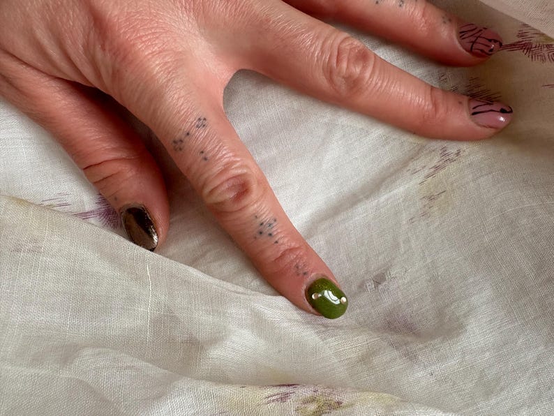 May include: Close-up of a hand with manicured nails. One nail is painted with a metallic bronze polish, and another is painted with a glossy olive green polish with a white dot. The other nails have a pink base with black lines.