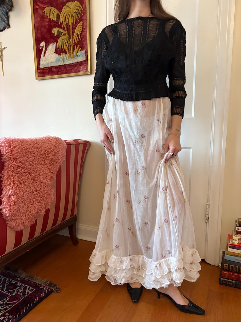 May include: A black lace long-sleeve top paired with a white floral print skirt. The skirt has a ruffled hem. The outfit is vintage-inspired and suitable for a special occasion or everyday wear.