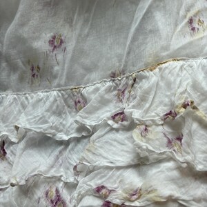May include: Close-up of a white fabric with a floral pattern. The fabric has a ruffled edge and is adorned with small, purple and yellow floral designs. The material appears to be lightweight and slightly translucent, suggesting a delicate texture.