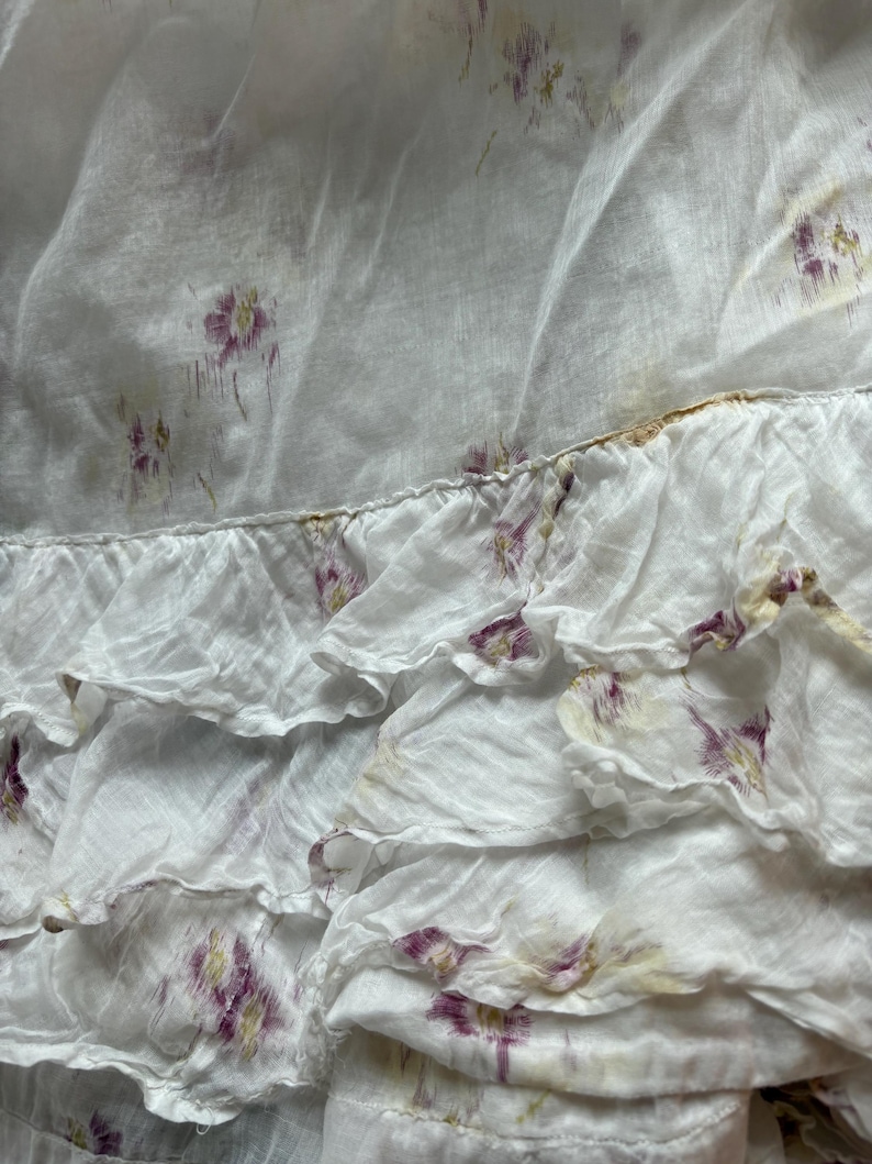 May include: Close-up of a white fabric with a floral pattern. The fabric has a ruffled edge and is adorned with small, purple and yellow floral designs. The material appears to be lightweight and slightly translucent, suggesting a delicate texture.