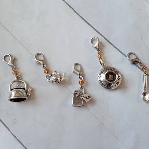 May include: Set of five silver charm clips with tea-themed designs. The charms include a teapot, a teacup, a tea bag, a kettle, and a spoon. The charms are attached to silver clips with orange beads.