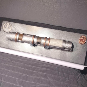 May include: A silver and copper lightsaber replica mounted on a silver plaque with a Rebel Alliance symbol and a copper symbol. The lightsaber is on a white display stand.