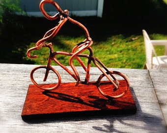 Op de afbeelding: Een sculptuur van koperdraad van een persoon die op een fiets rijdt op een houten basis. Het beeld is een uniek en grillig kunstwerk.