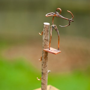 May include: A copper wire sculpture of an archer standing on a wooden platform. The platform is attached to a wooden stick with three copper wire hooks.